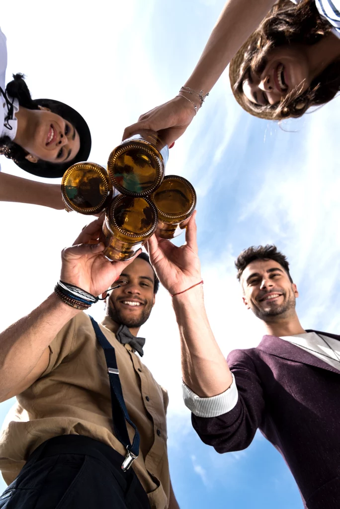 MÜW BEER : groupe d’amis qui trinquent avec des bouteilles de bière, vue en contre-plongée sous un ciel bleu.